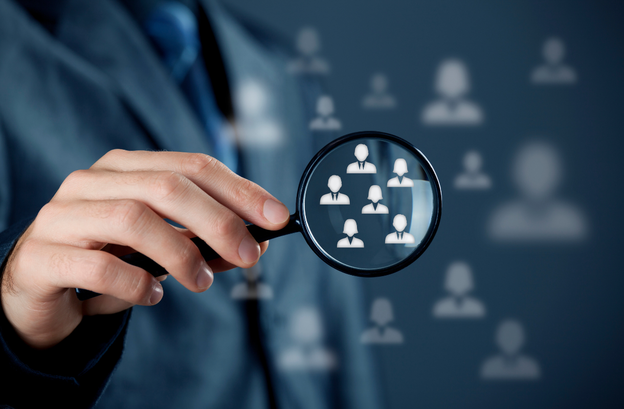 A person holds a magnifying glass focusing on icons of people, symbolizing analysis, recruitment, or searching for specific individuals within a group.