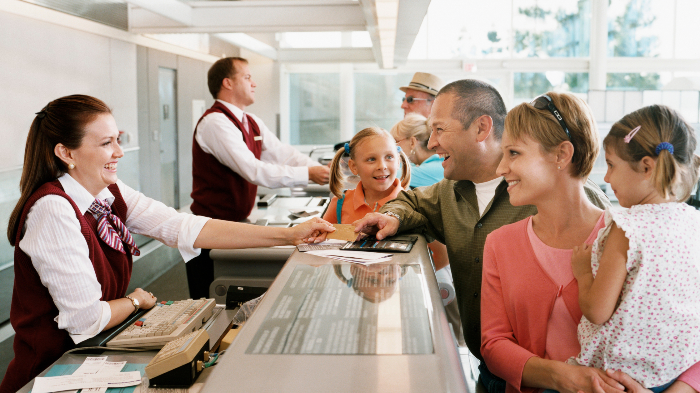 A family checks in at an airport counter for their group booking airline reservation, handing documents to a smiling employee while other travelers are assisted in the background.